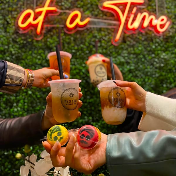 Four hands holding PYO Chai boba drinks and two macarons against a green wall with neon sign.