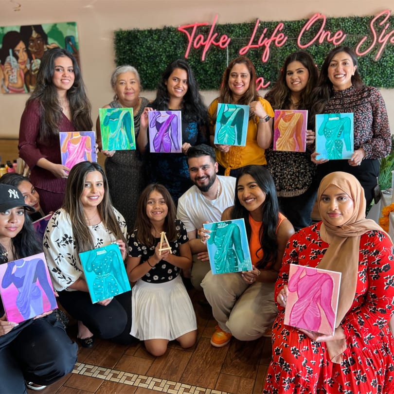 Diverse group of people smiling and holding up colorful paintings of saris at PYO Chai.