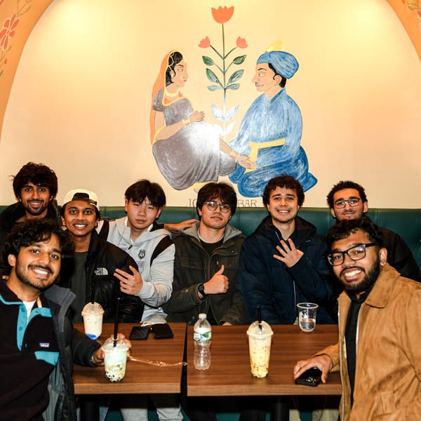 Eight young men smiling at PYO Chai, with boba drinks on tables and a mural behind them.