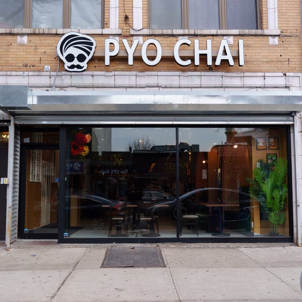 Exterior of PYO CHAI boba shop with a turbaned logo, large windows, and visible interior seating.