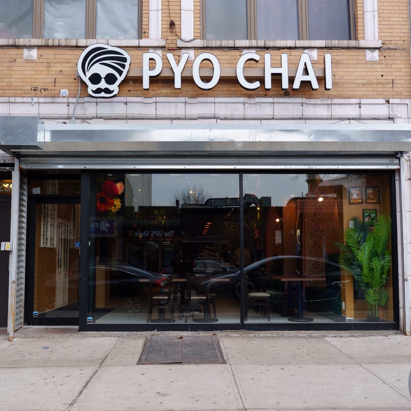 Exterior of PYO CHAI boba shop with a turbaned logo, large windows, and visible interior seating.