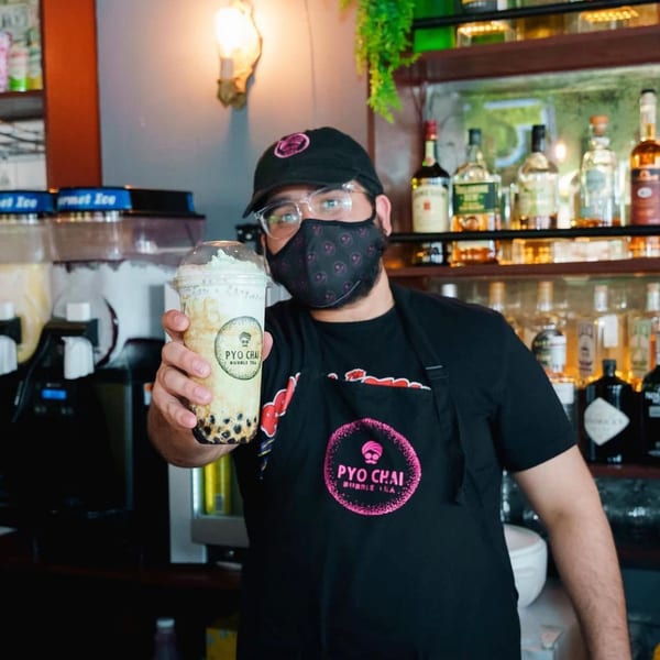 PYO Chai employee in a mask and apron holding up a boba drink with whipped cream.