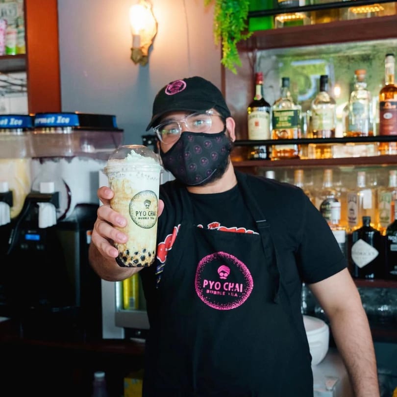 PYO Chai employee in a mask and apron holding up a boba drink with whipped cream.