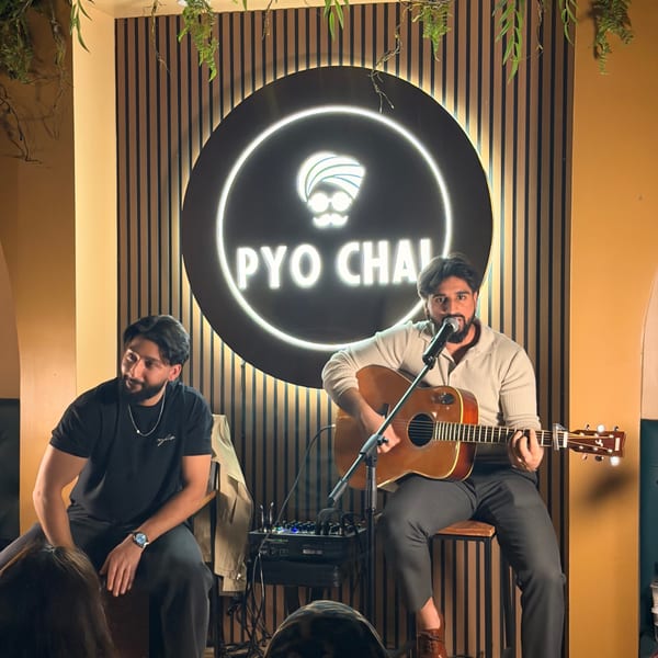 Two men performing music, one singing and playing guitar, in front of a glowing PYO CHAI sign.