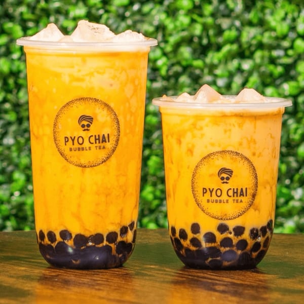 Two PYO Chai bubble teas with ice and dark boba pearls, on a wooden table with green foliage.